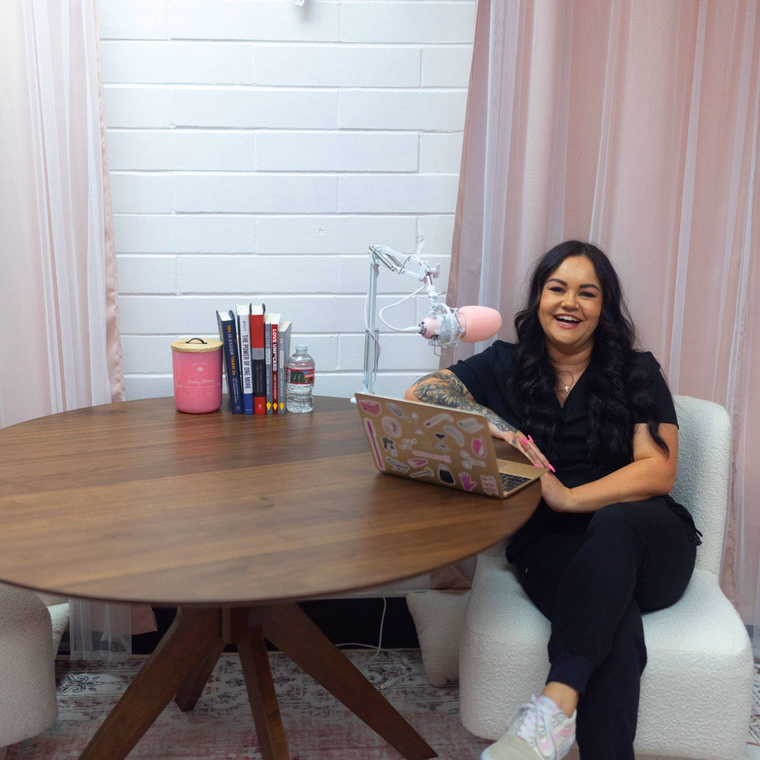 Photo of Bree owner of Lash and Wax OC smiling and sitting at a table in front of her laptop and microphone.